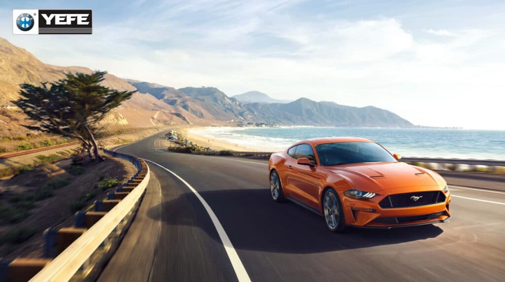 An orange Ford Mustang speeding along a scenic coastal highway, representing steady-speed driving conditions that are ideal for engine oil performance. This image contrasts with stop-and-go city driving, highlighting how consistent engine temperatures and reduced thermal cycling allow oil to maintain optimal viscosity and protection. Perfect for illustrating the benefits of using high-quality lubricants like YEFE in diverse driving environments.