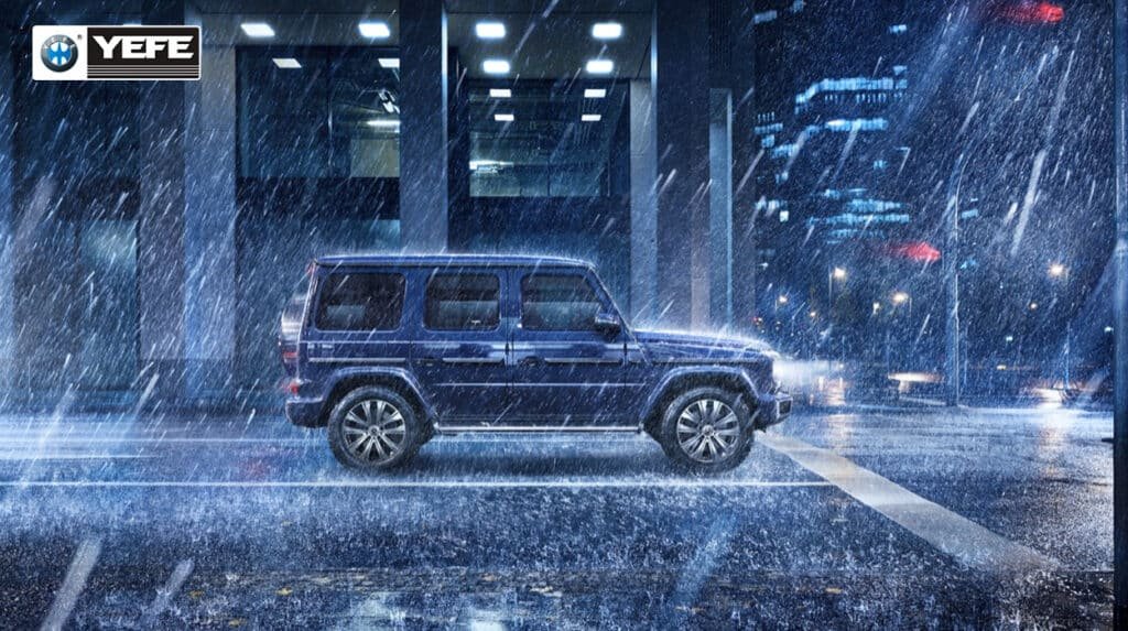 A dark blue Mercedes-Benz G-Class SUV is shown driving through heavy rain at night in an urban environment, with water splashing around its wheels and city lights reflecting on the wet pavement. The vehicle's iconic boxy silhouette, side-opening tailgate, and spare tire with the Mercedes star emblem are clearly visible. This image illustrates the G-Class's robust all-weather performance, leveraging its full-time four-wheel drive, three 100% differential locks, and high ground clearance to maintain traction and control in challenging conditions. The scene underscores the vehicle's dual identity as both a luxury icon and a capable off-roader, engineered to deliver confidence and safety regardless of weather or terrain.